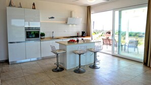 A modern kitchen with white cabinetry, a central island with bar stools, a built-in oven, and a large sliding glass door leading to an outdoor patio. - Complesso Villa Vanissia (Castrignano del Capo)