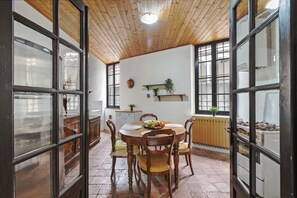 A dining area with a round wooden table, chairs, and a bowl of fruit. There are windows with wooden frames and a ceiling with wooden beams.