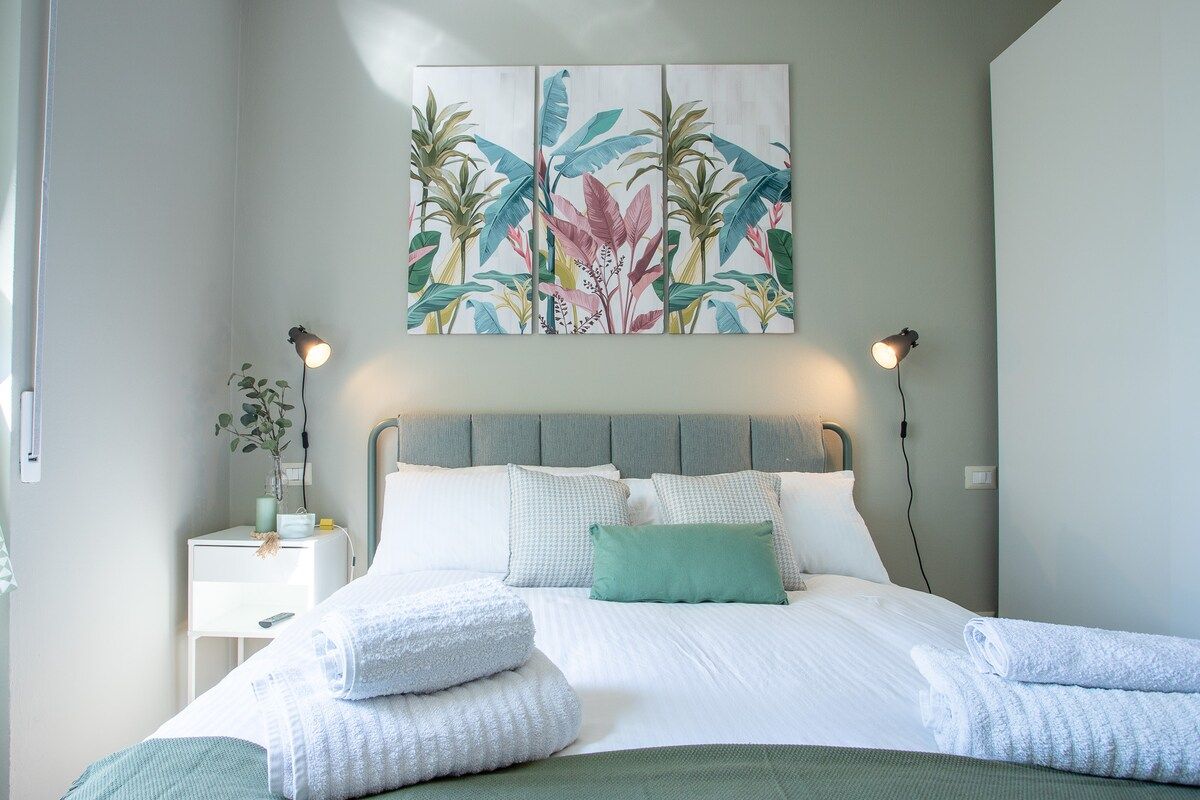 Un lit bien fait, avec du linge de lit blanc et un plaid vert, un tableau représentant des feuilles tropicales accroché au mur, et des lampes de chevet.