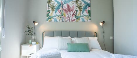 A neatly made bed with white linens and a green throw, a wall-mounted painting of tropical leaves, and bedside lamps.