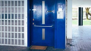 Una puerta azul con manija metálica y teclado de seguridad, una reja metálica a la izquierda y piso de ladrillo.