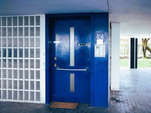 A blue door with a metal handle and a security keypad, a metal grid on the left, and a brick floor. - Porta21 Studio [Linate, Osp.Monzino, Arena Santa Giulia] (Milano)