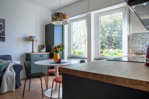A modern kitchen with a central island, a dining area with a table and chairs, and a view of greenery outside.