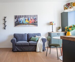 A modern living room with a grey sofa, a green chair, a cityscape painting, and a dining area with a table and chairs. - Porta21 Studio [Linate, Osp.Monzino, Arena Santa Giulia] (Milano)