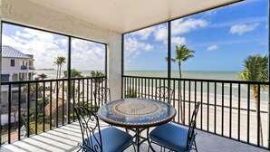 A balcony with a round table and chairs, overlooking a beach with palm trees and a clear sky. - UPDATED 2B/2B BEACHFRONT CONDO SEASIDE 301 (Fort Myers Beach)