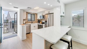 A modern kitchen with white cabinetry, a central island, and a large window with blinds. - UPDATED 2B/2B BEACHFRONT CONDO SEASIDE 301 (Fort Myers Beach)