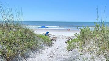 A sandy beach path leading to the ocean with a blue umbrella and two beach chairs.