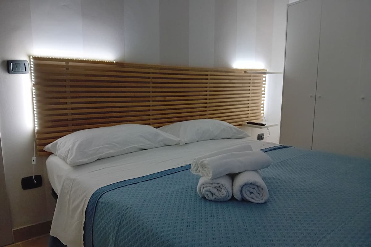 Uma cama bem arrumada com lençóis brancos e um cobertor azul, uma cabeceira de madeira e uma mesa de cabeceira com um controle remoto.