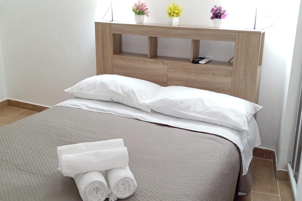 Cama bem arrumada com lençóis brancos e toalhas dobradas, cabeceira de madeira e uma pequena prateleira com plantas em vasos.