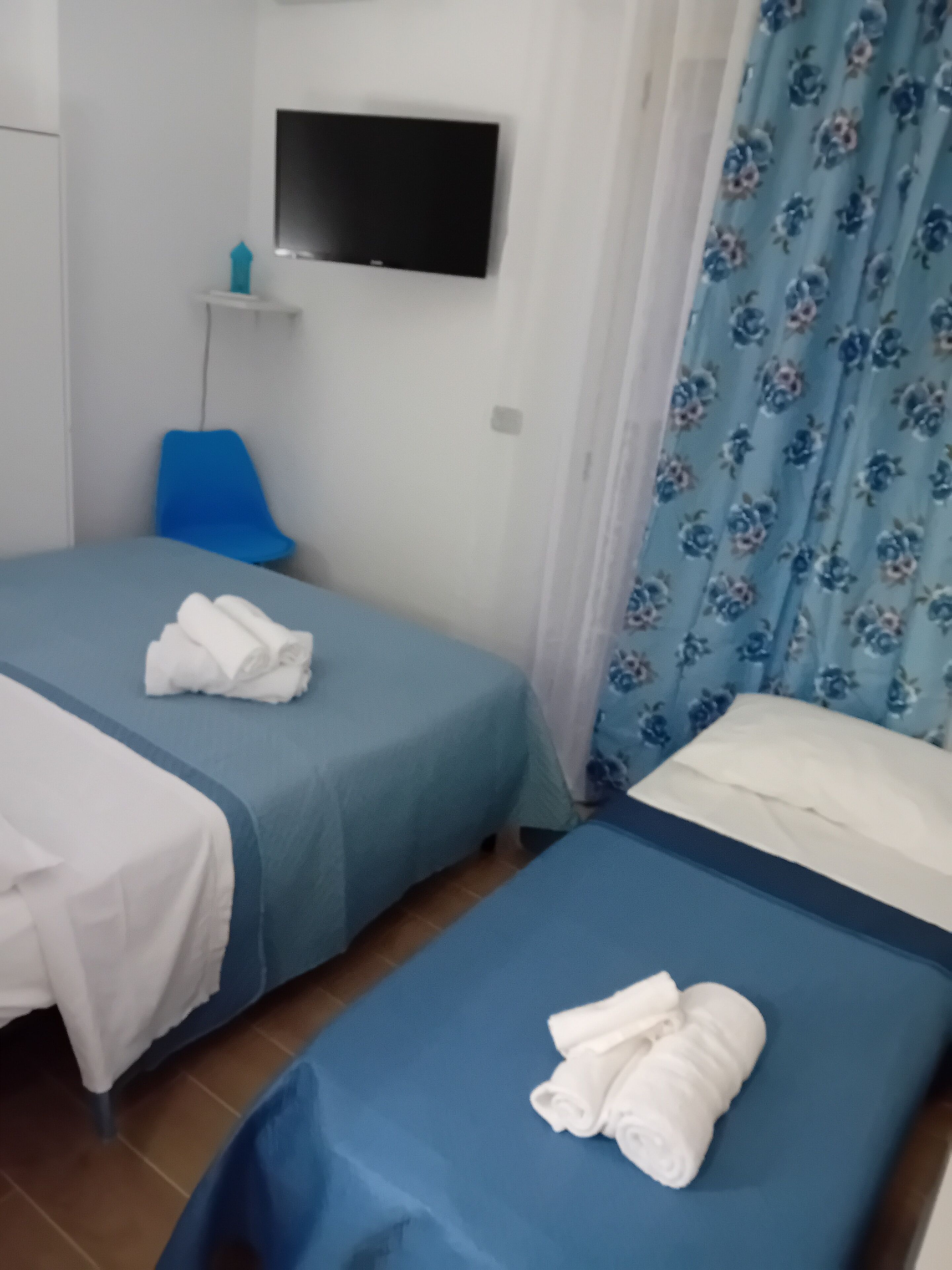 A hotel room with two single beds, a TV mounted on the wall, a blue chair, and a floral curtain.