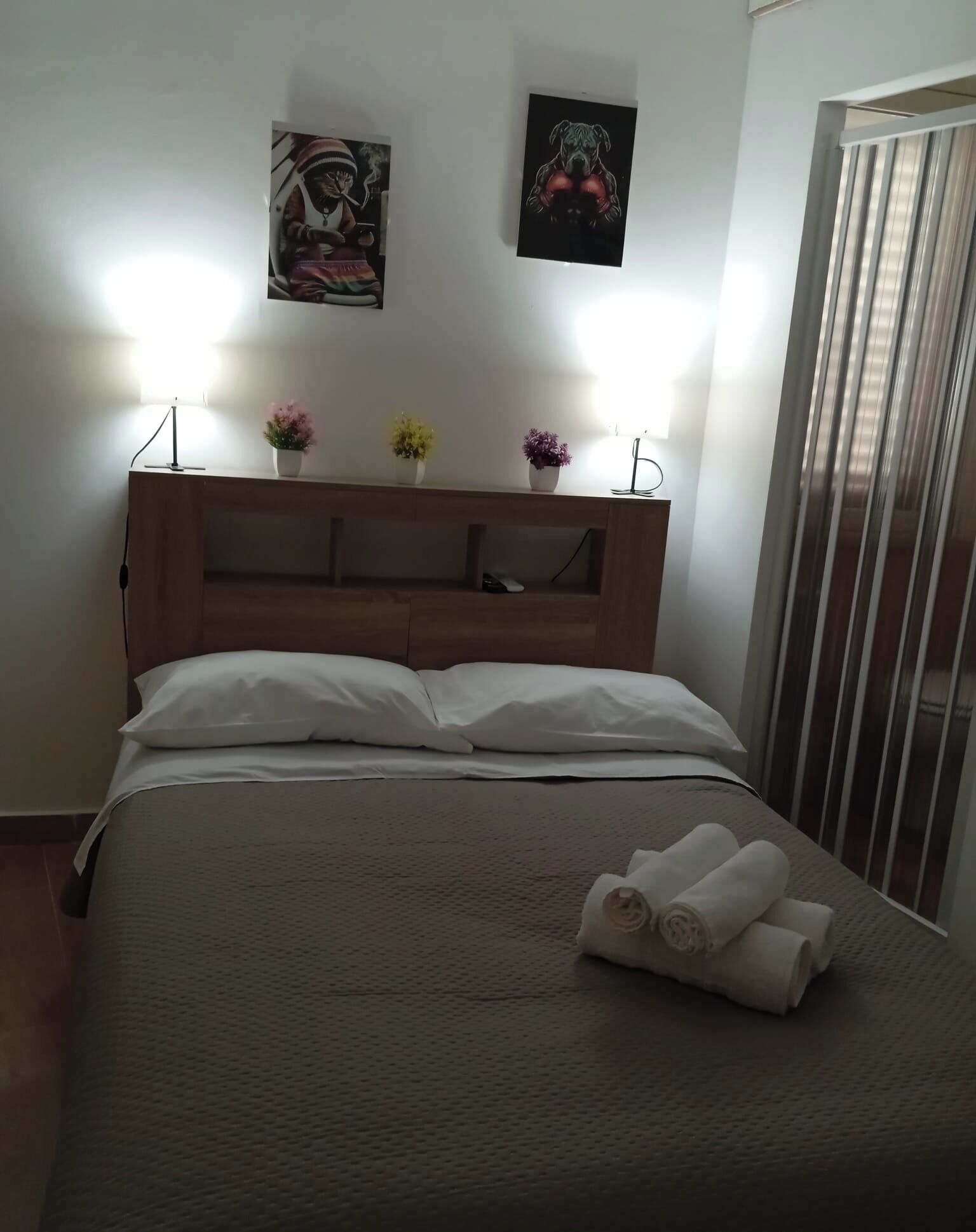 Hotelkamer met houten hoofdbord, twee kunstwerken aan de muur, een bedlamp, een bed met witte lakens en een raam met zonwering.