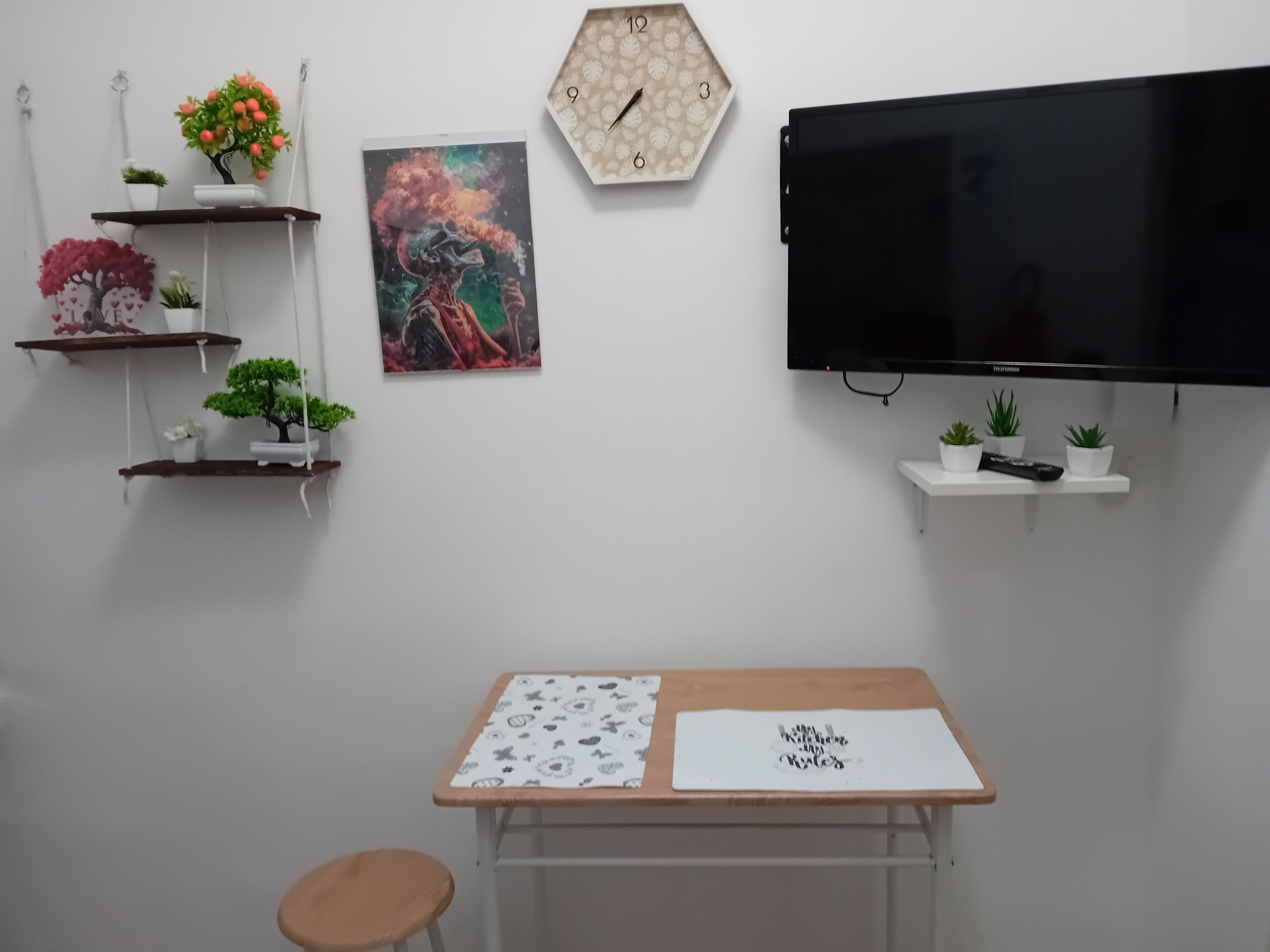 Una stanza con una TV a schermo piatto, un tavolo in legno con tovaglietta bianca, uno sgabello e mensole a parete con piante e un orologio.