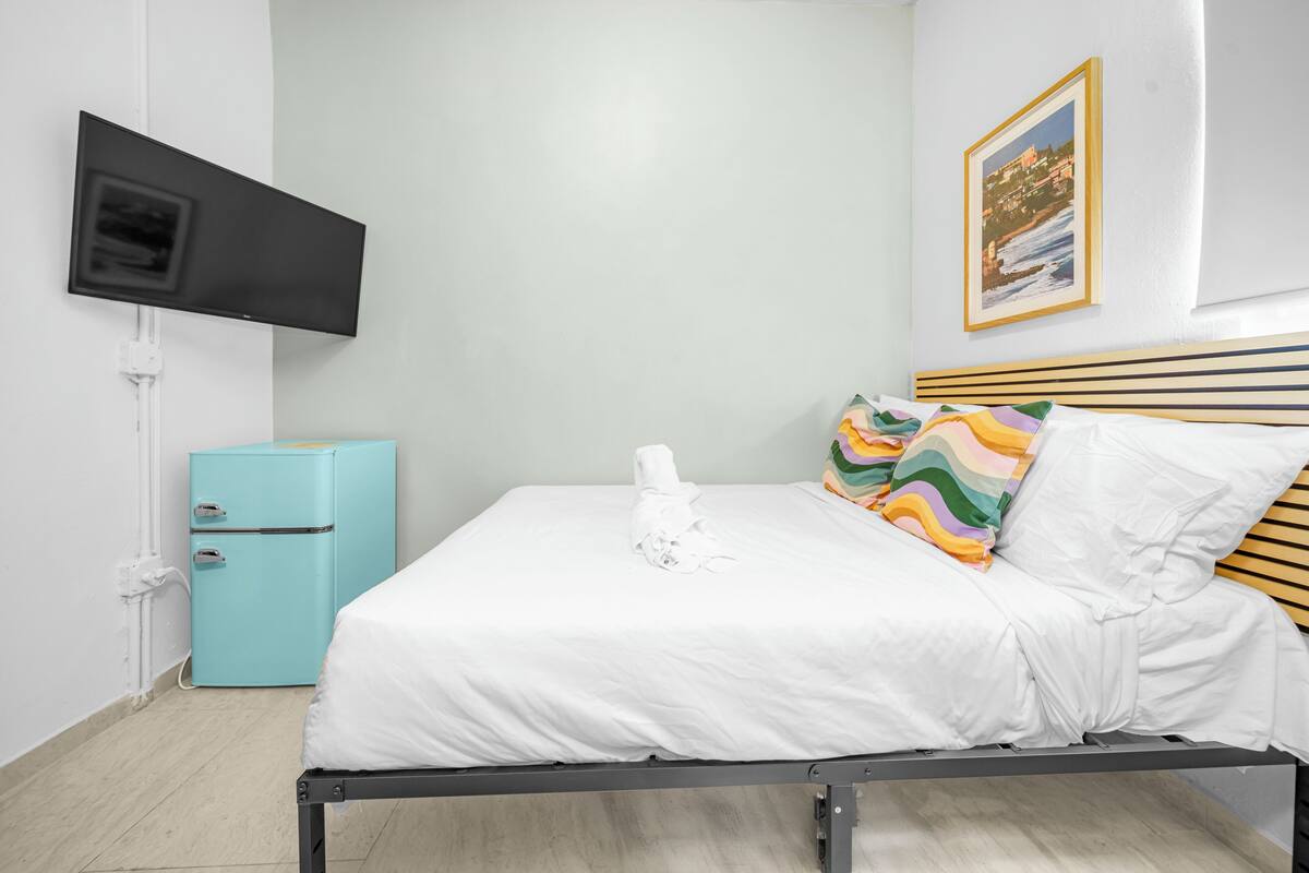A neatly made bed with white linens, a teal-colored mini-fridge, a flat-screen TV mounted on the wall, and a framed picture above the bed.