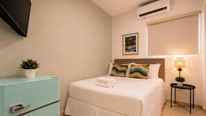 Hotelkamer met een bed, een bedlamp, een tafeltje met een plant, een tv aan de muur en een raam met zonwering.