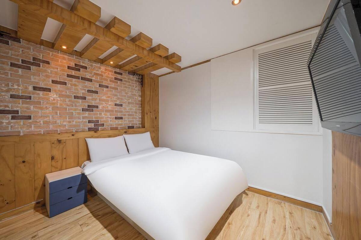 A bedroom with a brick wall, wooden ceiling beams, a bed, a nightstand, and a window with blinds.