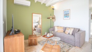 A living room with a grey sofa, wooden side table, and a mirror reflecting the kitchen area. - Houat (Le Diamant)