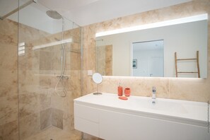 A modern bathroom with a glass-enclosed shower, a white vanity with a mirror, and a beige tiled wall. - Houat (Le Diamant)