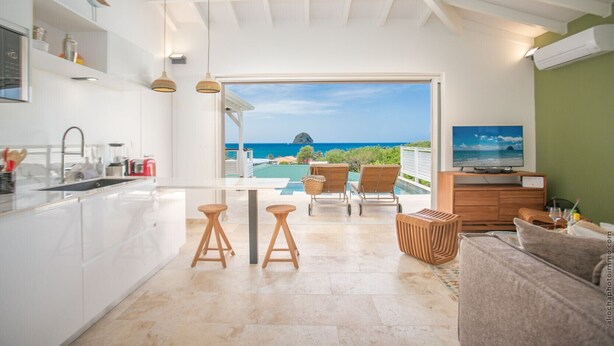 A modern kitchen with a view of the ocean, a dining area with stools, and a TV on a wooden cabinet. - Houat (Le Diamant)