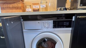 Una lavadora de carga frontal con panel de control, un panera de madera y botes etiquetados como 'café' y 'azúcar morena' encima.