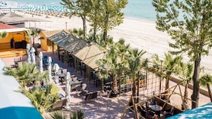 Et udendørs opholdsområde med borde og parasoller med udsigt over en strand med parasoller og mennesker.