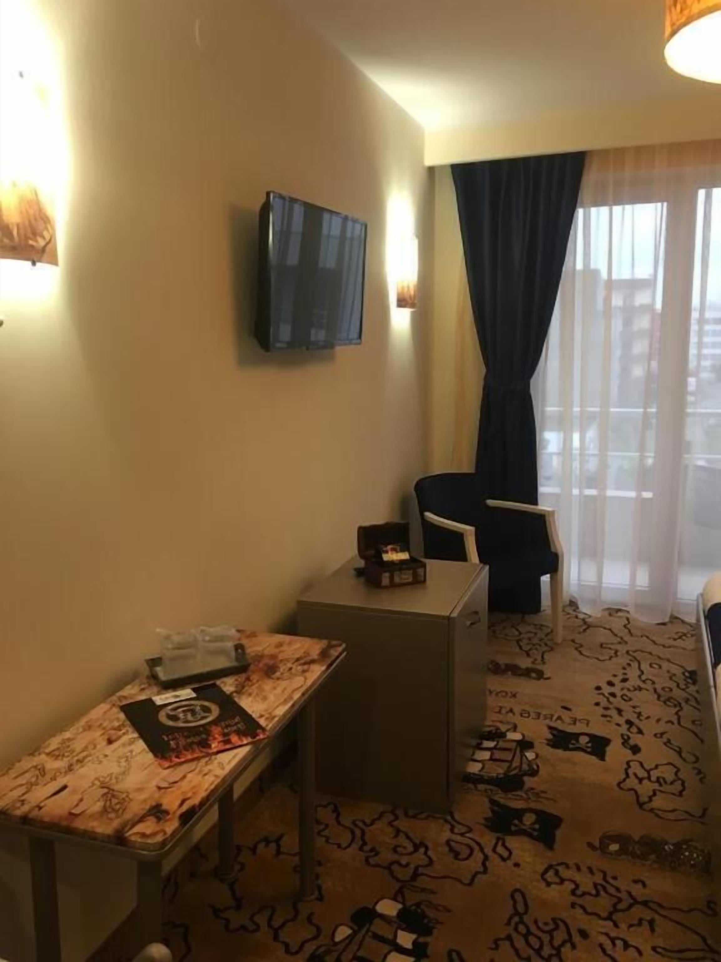 A hotel room with a small desk, a chair, a television mounted on the wall, and a view of the city through the window.