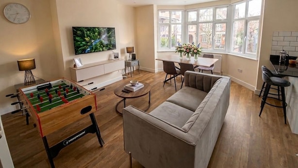 A living room with a foosball table, a sofa, a dining table with chairs, a TV, and a window with a view. - Lovely Two Bed Apartment In London (South Croydon)