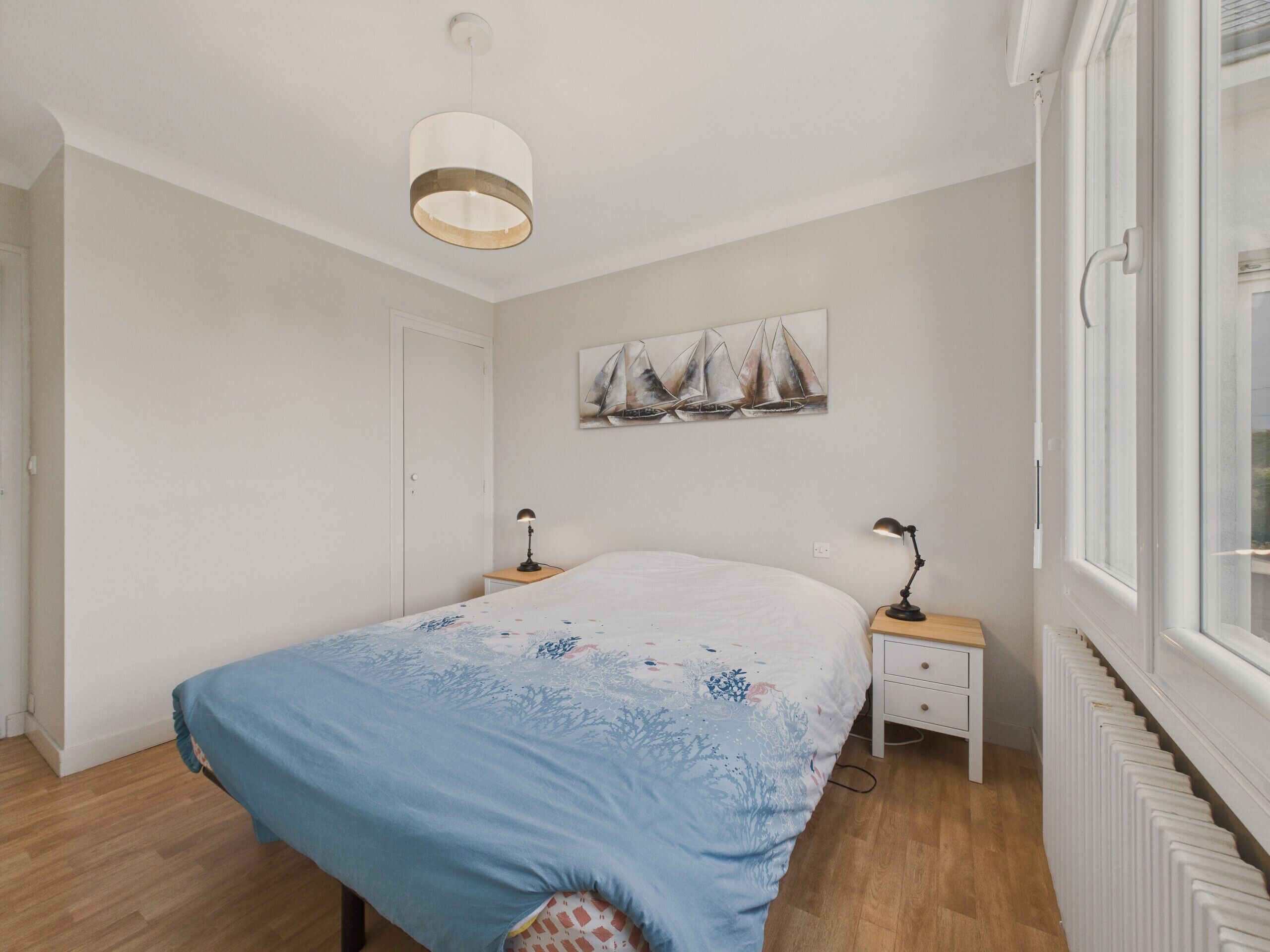 A bedroom with a bed, two bedside tables, a ceiling light, a window, and a radiator.