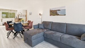 A modern living room with a grey sectional sofa, a dining area with a round table and chairs, and a coastal landscape painting on the wall. - Sea view in the city center - Apartment for 6 (Erquy)