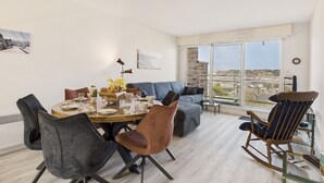 A modern living room with a dining area, a sofa, a rocking chair, and a large window with a city view. - Sea view in the city center - Apartment for 6 (Erquy)