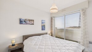 A bedroom with a large bed, a bedside table with a lamp, a painting on the wall, and a view of the sea through a large window. - Sea view in the city center - Apartment for 6 (Erquy)