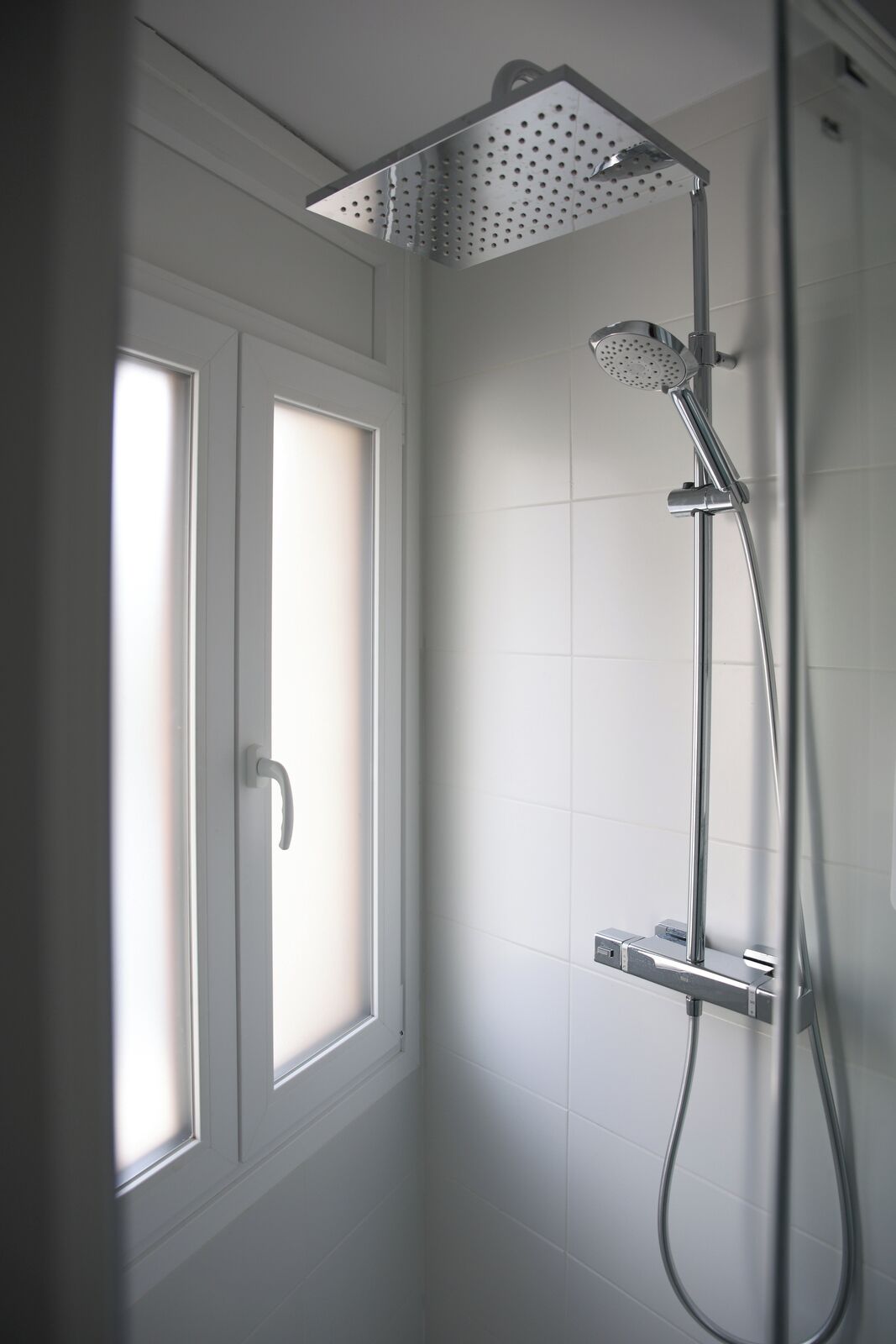 A shower with a handheld showerhead and a wall-mounted faucet, a window with a frosted glass door, and white tiled walls.