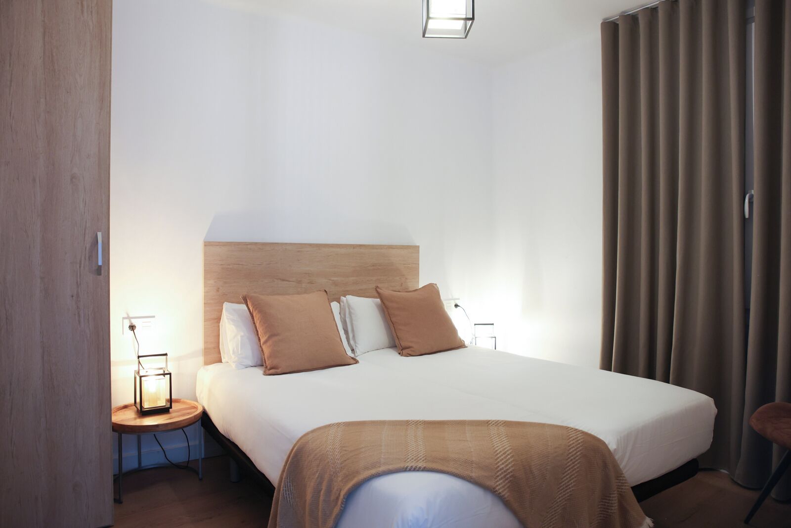A hotel room with a large bed, a wooden headboard, a bedside table with a lamp, and a window with curtains.