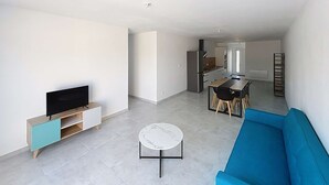 Ein großzügiger Wohnbereich mit Flachbildfernseher, einem blauen Sofa, einem Esstisch mit Stühlen und einer Küche im Hintergrund.