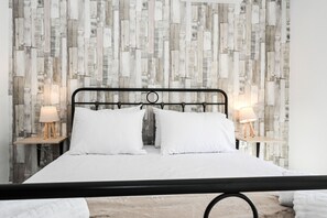 A neatly made bed with white linens and two pillows, flanked by bedside tables with lamps, set against a wall with a wooden plank pattern. - Casa 30 House Complex Center of Ialysos (Rhodes)