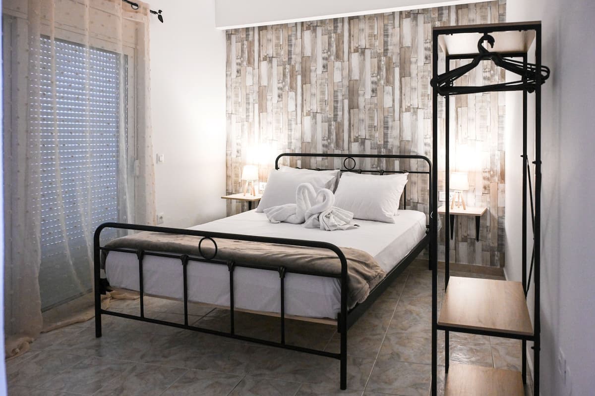 Ein ordentlich bezogenes Bett mit einem herzförmigen Handtuch, ein Nachttisch mit Lampe und eine Wand mit rustikaler Holzoptik.