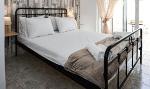 A neatly made bed with white linens and a brown throw blanket, a bedside table with a lamp, and a view of the outdoors through a window with curtains. - Casa 30 House Complex Center of Ialysos (Rhodes)
