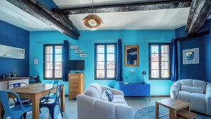 A modern living room with a blue color scheme, featuring a sofa, chairs, a wooden dining table, and a TV.