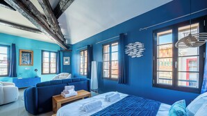 A hotel room with a large bed, a blue sofa, a wooden nightstand, and a view of the outside through the windows.