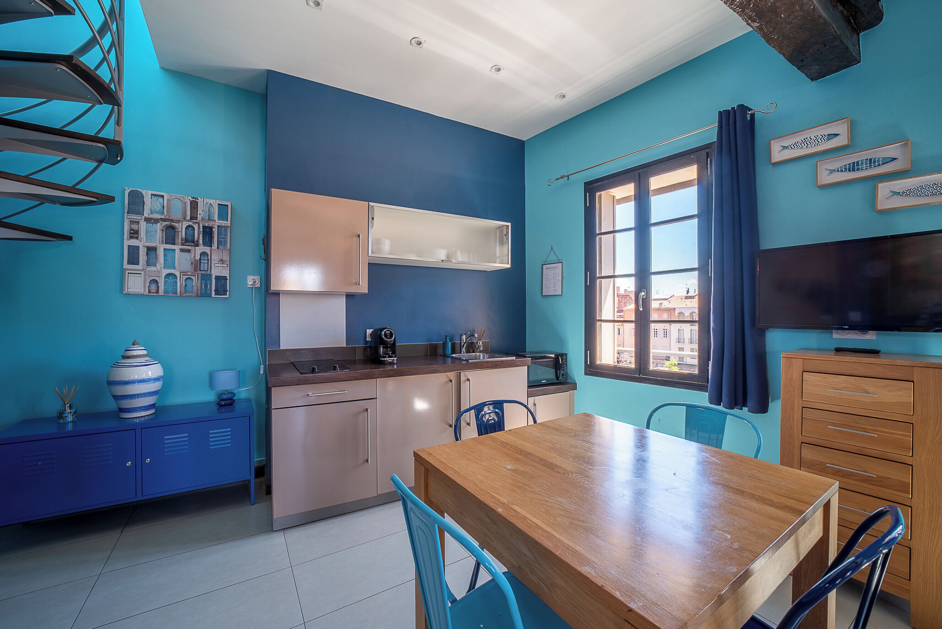 A compact kitchen with a wooden table, blue chairs, and a window with curtains.