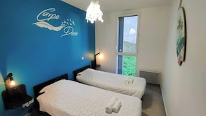 A hotel room with two beds, a blue accent wall, a chandelier, and a window with a view of greenery. - Apartment with Pool, Air Conditioning, WiFi, and Garage (Collioure)
