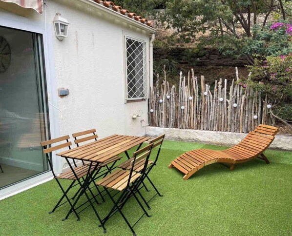 A patio with a wooden table and chairs, a sun lounger, and a small white building with a window and a lantern. - Charming Collioure • Terrace • Parking • Wi-Fi (Collioure)