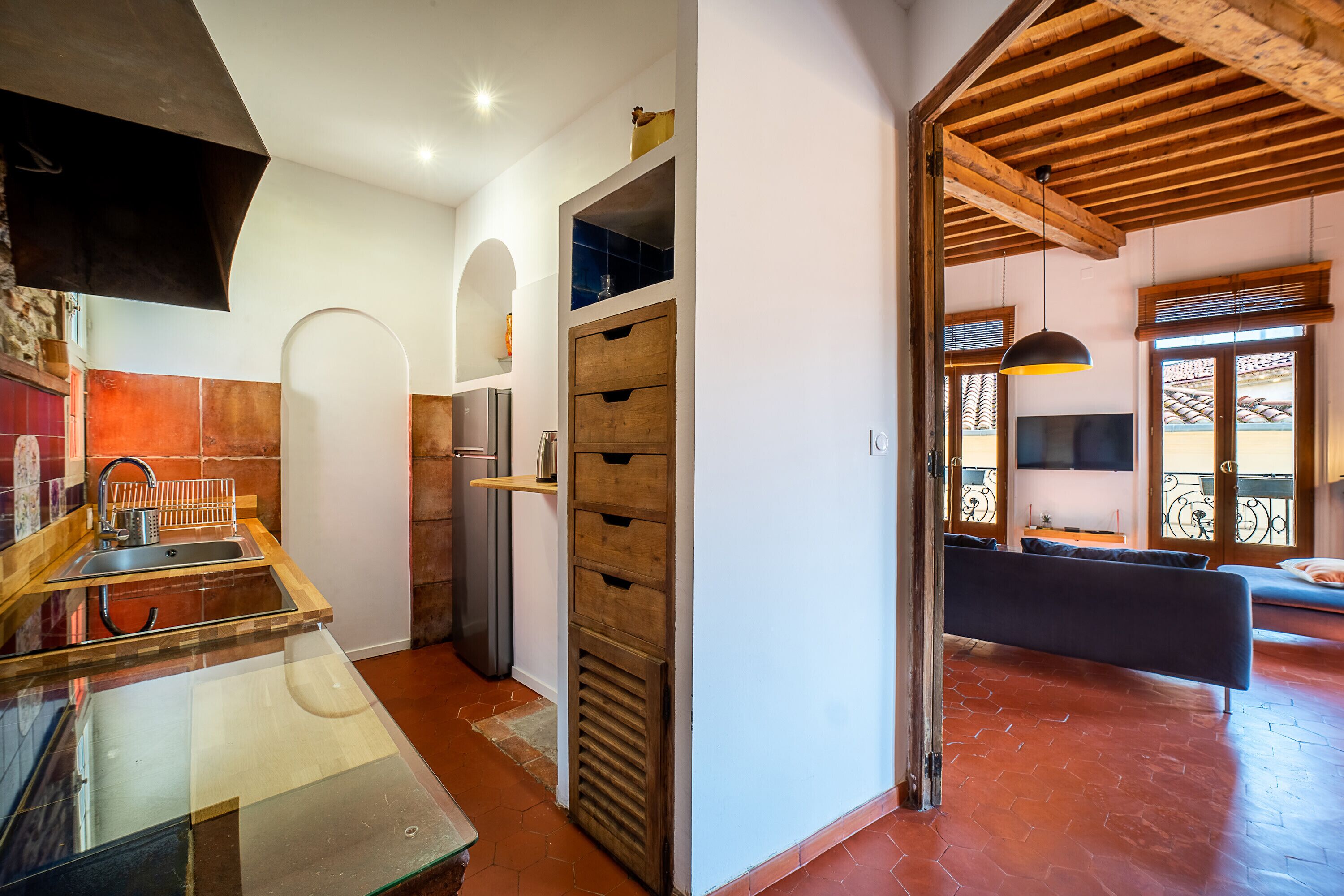A modern kitchen with a glass countertop, stainless steel sink, and wooden cabinets. There is a dining area with a sofa and a TV.