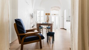 Una habitación amplia con una mesa de comedor de madera, sillas y un taburete pequeño. Se observa un radiador a la izquierda y una zona de cocina al fondo.