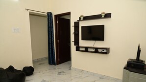 Una stanza con una TV fissata al muro, un piccolo ripiano con oggetti decorativi, un pouf nero e un mobile contenente una stampante.