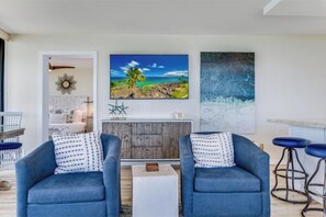 Een woonkamer met twee blauwe fauteuils, een witte salontafel, een houten kast en een groot schilderij van een tropisch landschap.