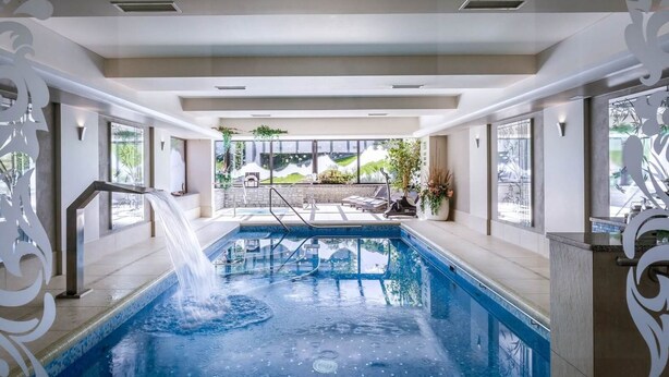 An indoor swimming pool with a waterfall feature, surrounded by lounge chairs and a view of a landscaped garden. - Rezydencja nad Wodospadem Spa&Wellness (Jelenia Gora)