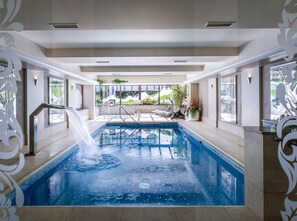 An indoor swimming pool with a waterfall feature, surrounded by lounge chairs and a view of a landscaped garden. - Rezydencja nad Wodospadem Spa&Wellness (Jelenia Gora)