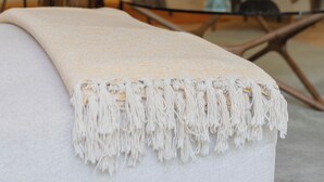 Una sala de estar moderna con una mesa de centro de vidrio, un sofá blanco con un plaid con flecos, y un comedor al fondo.