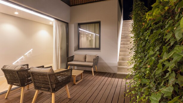 Un área de estar al aire libre con sillas de mimbre y una mesa, una ventana con cortinas y una escalera que conduce a un nivel superior.