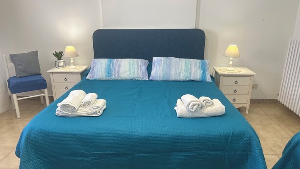 A neatly made bed with a teal bedspread, two pillows, and two rolled towels. There are two bedside tables with lamps, a chair, and a small plant. - Cozy house with veranda (Otranto)